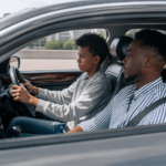 A father sits beside his son who is learning to drive, inside a car.